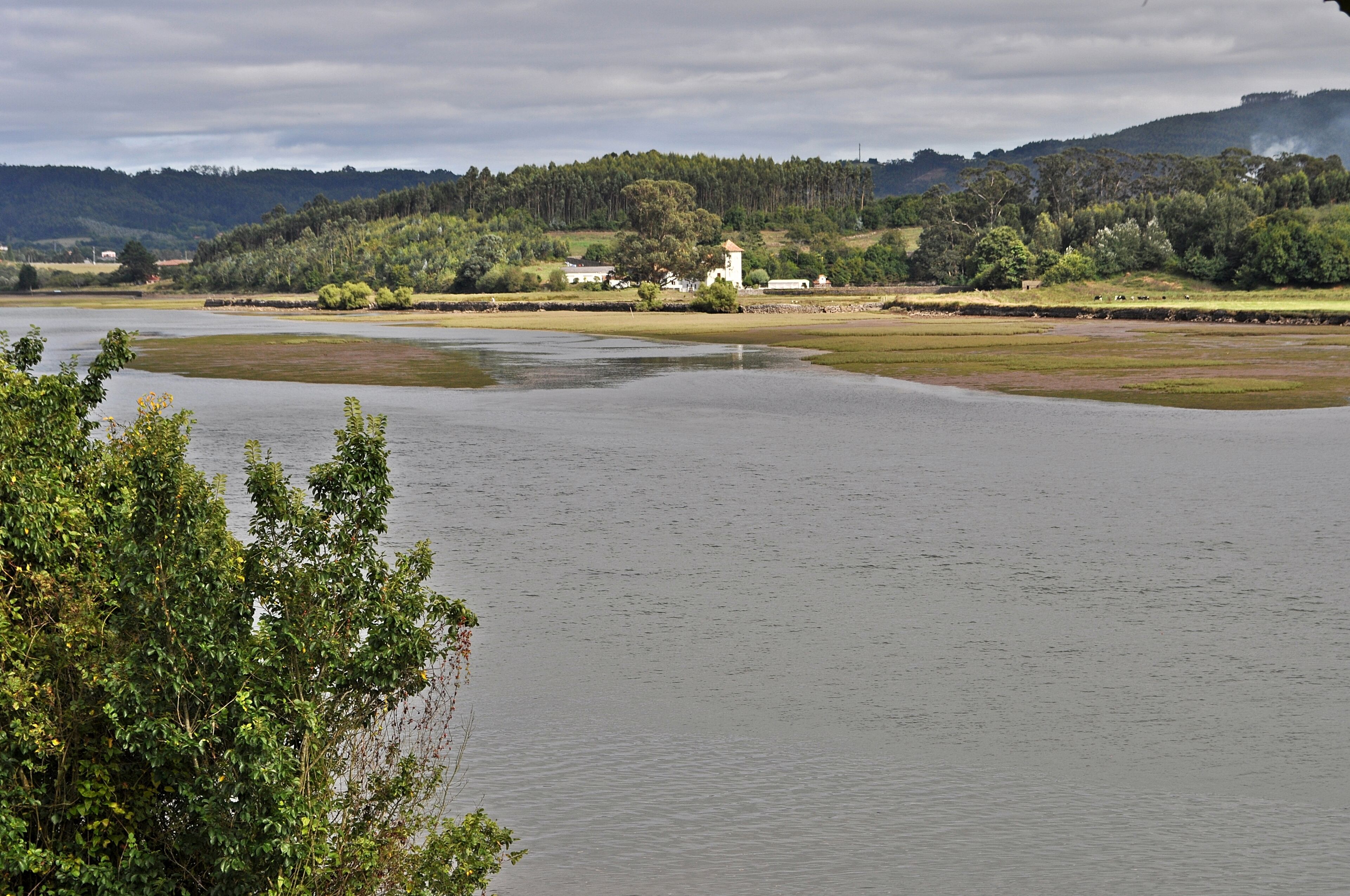 A calm lake with a white building on the shore, surrounded by greenery and hills.