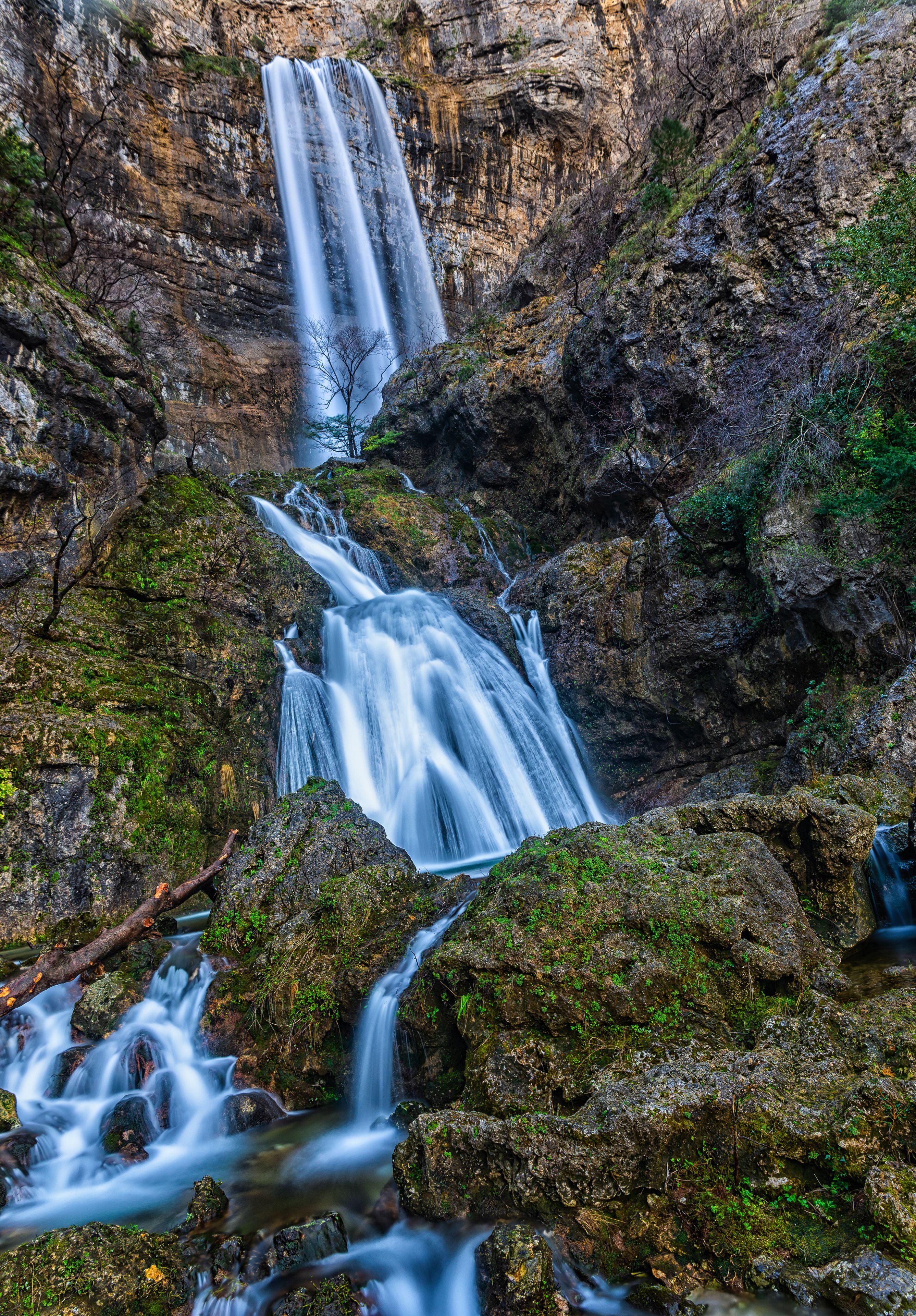 A multi-tiered waterfall cascading over rocky terrain with moss-covered rocks and sparse vegetation.