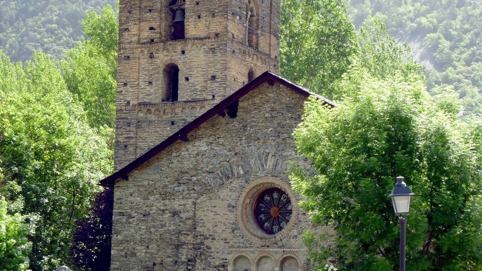 A stone church with a bell tower and a circular window, surrounded by trees and a mountain in the background.