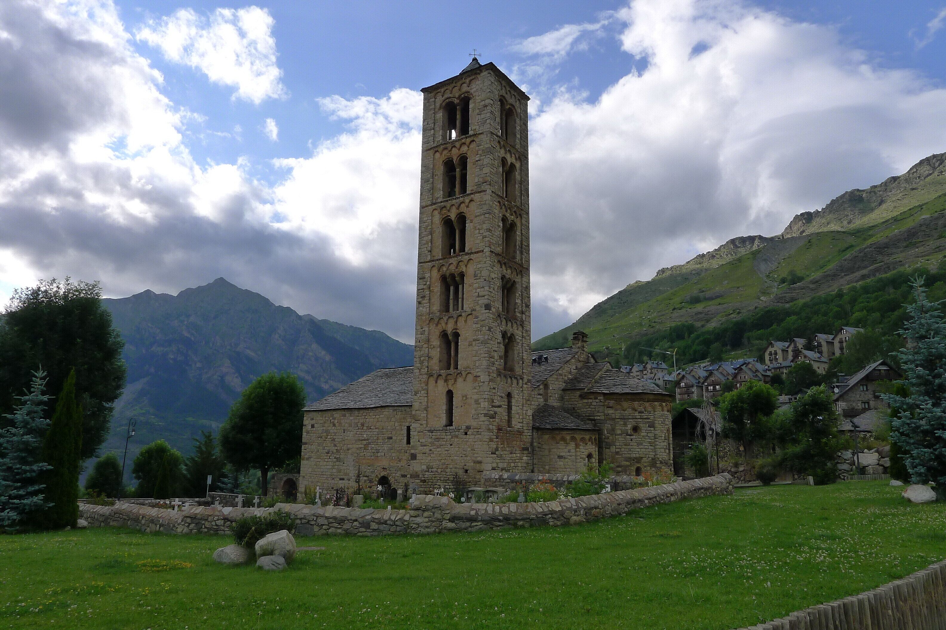 A stone church with a bell tower, surrounded by a grassy field and mountains in the background.
