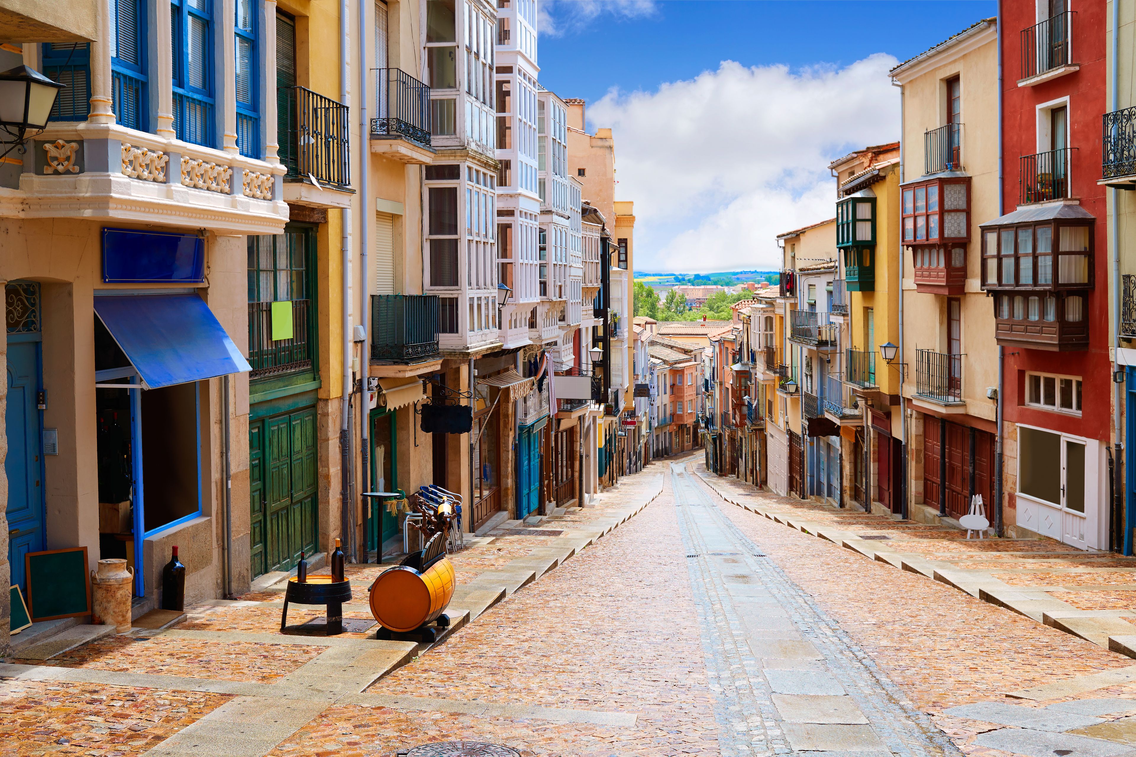 A cobblestone street lined with colorful buildings, a yellow barrel, and a blue awning.