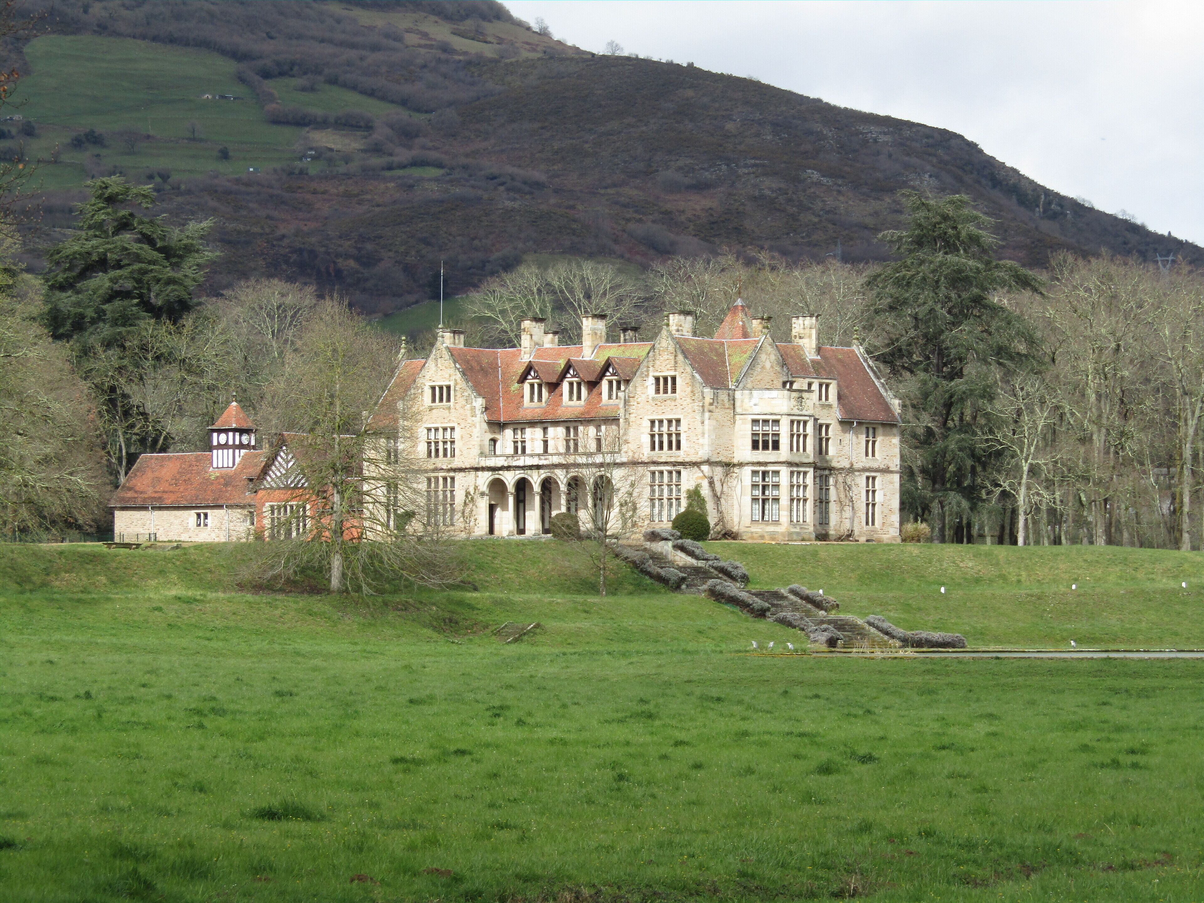A large stone manor house with a red-tiled roof, surrounded by a green field and a forested hill in the background.