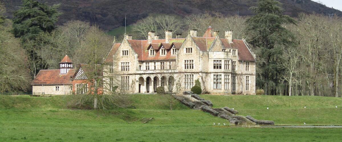 A large stone manor house with a red-tiled roof, surrounded by a green field and a forested hill in the background.