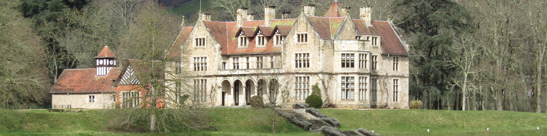 A large stone manor house with a red-tiled roof, surrounded by a green field and a forested hill in the background.
