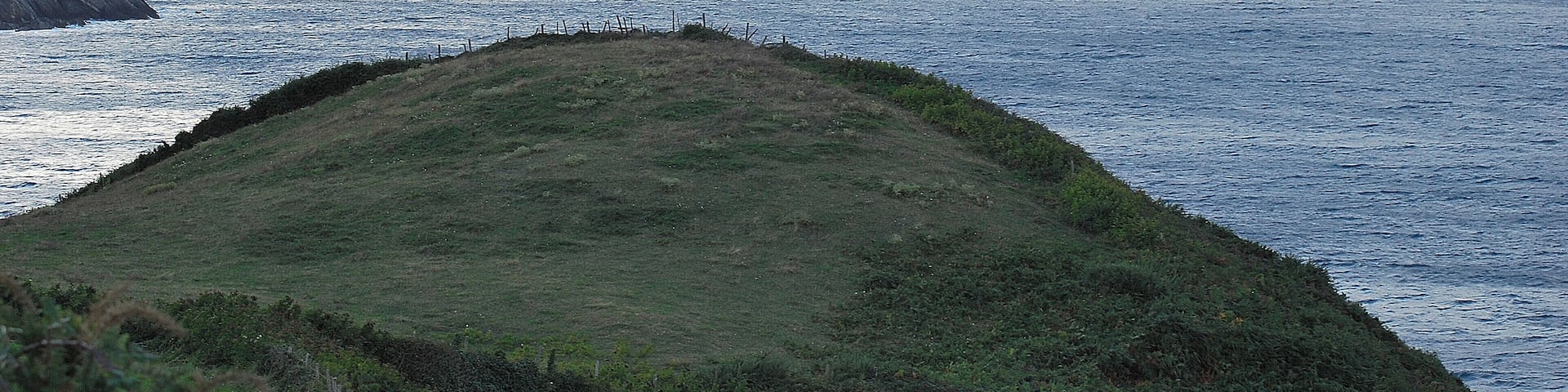 A coastal landscape with a grassy hill, cliffs, and a view of the sea.