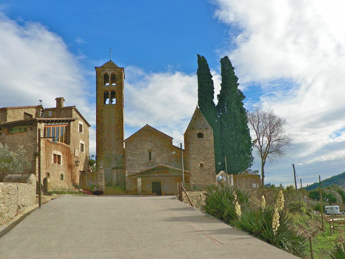 A stone church with a bell tower, surrounded by stone buildings and tall cypress trees.