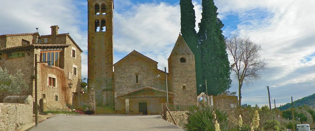 A stone church with a bell tower, surrounded by stone buildings and tall cypress trees.