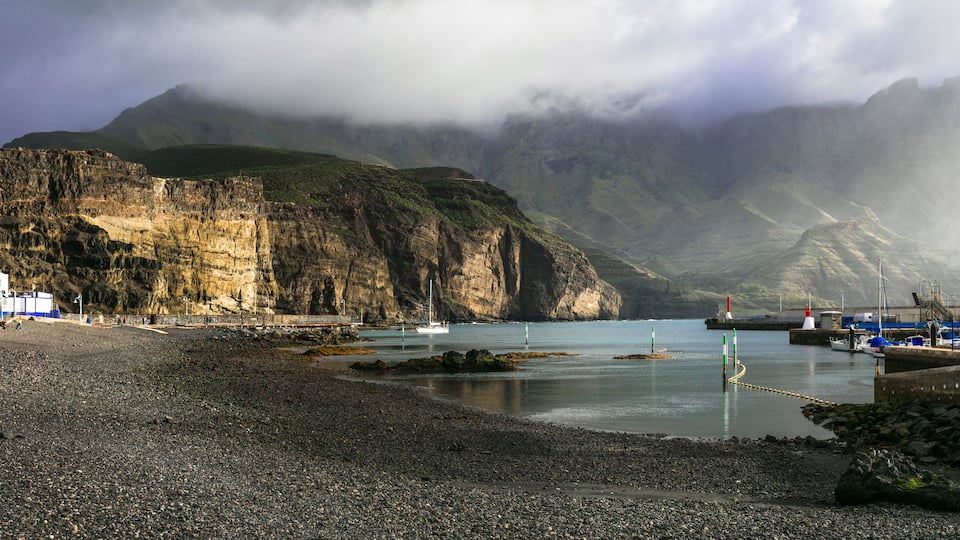 A rocky beach with a lighthouse, boats, and a mountainous backdrop.