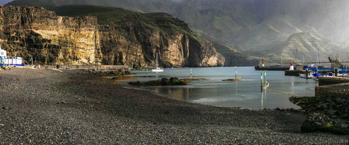A rocky beach with a lighthouse, boats, and a mountainous backdrop.
