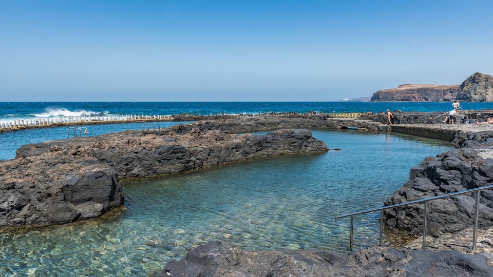 A rocky coastal area with clear water, a rocky breakwater, and a distant cliff.