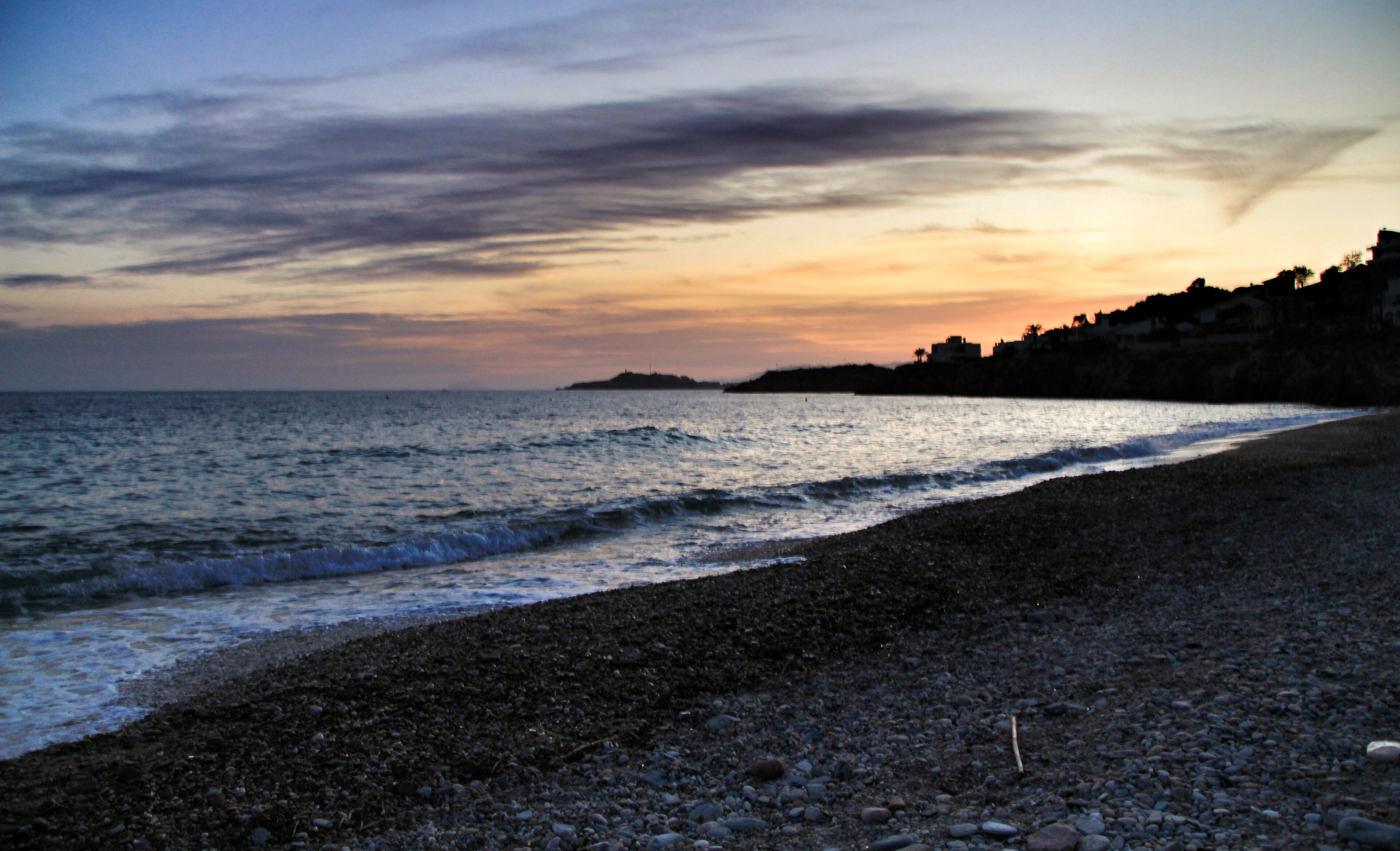 A pebbly beach with waves at sunset, a distant island, and a coastline with buildings.