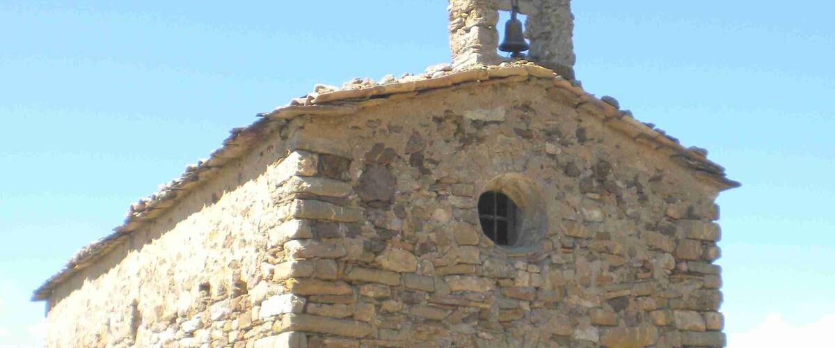 A small stone church with a bell tower and a cross on top, surrounded by a grassy field and mountains in the background.