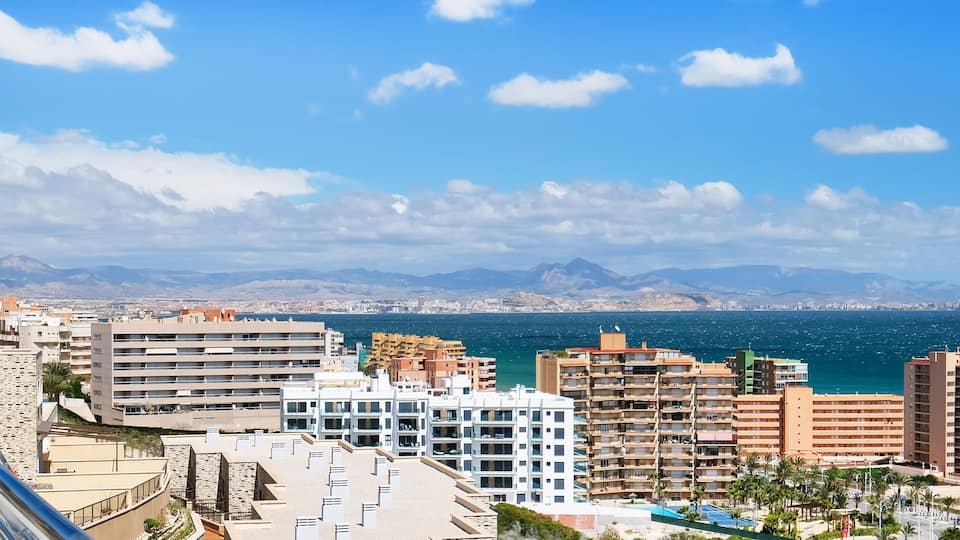 A coastal cityscape with modern buildings, a beach, and mountains in the background.