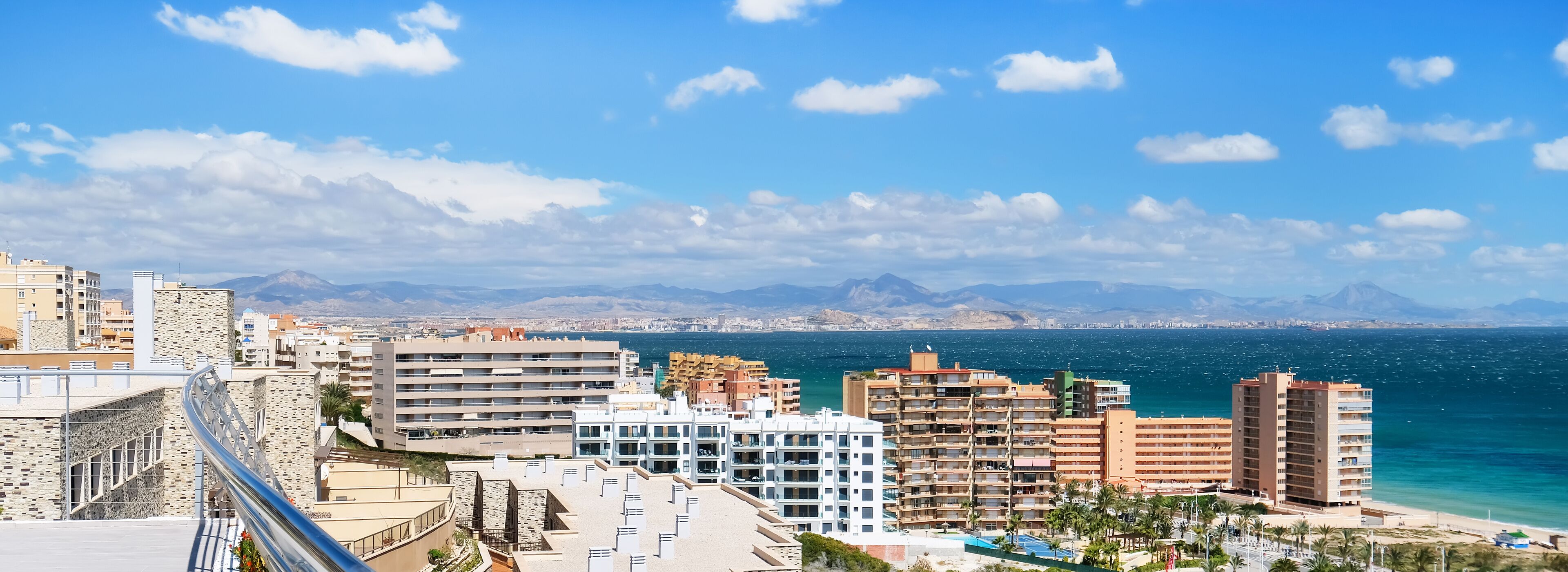 A coastal cityscape with modern buildings, a beach, and mountains in the background.