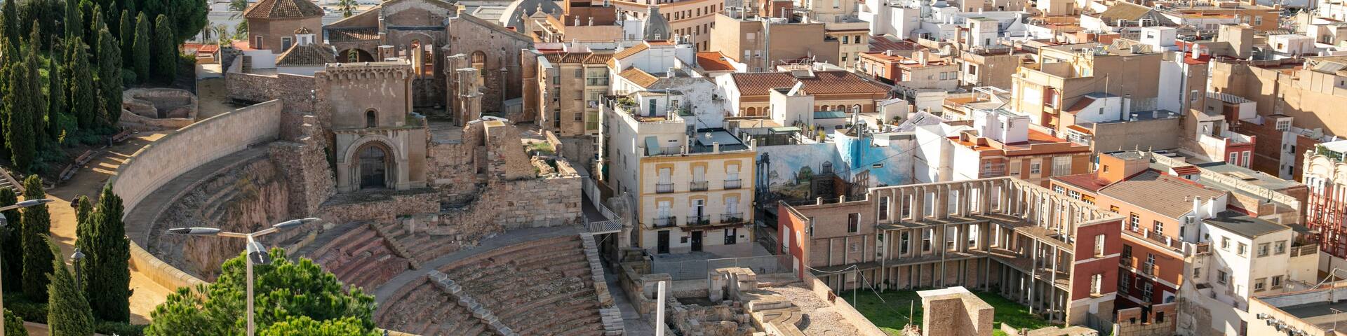 A coastal city with a historic amphitheater, modern buildings, and a mountainous backdrop.