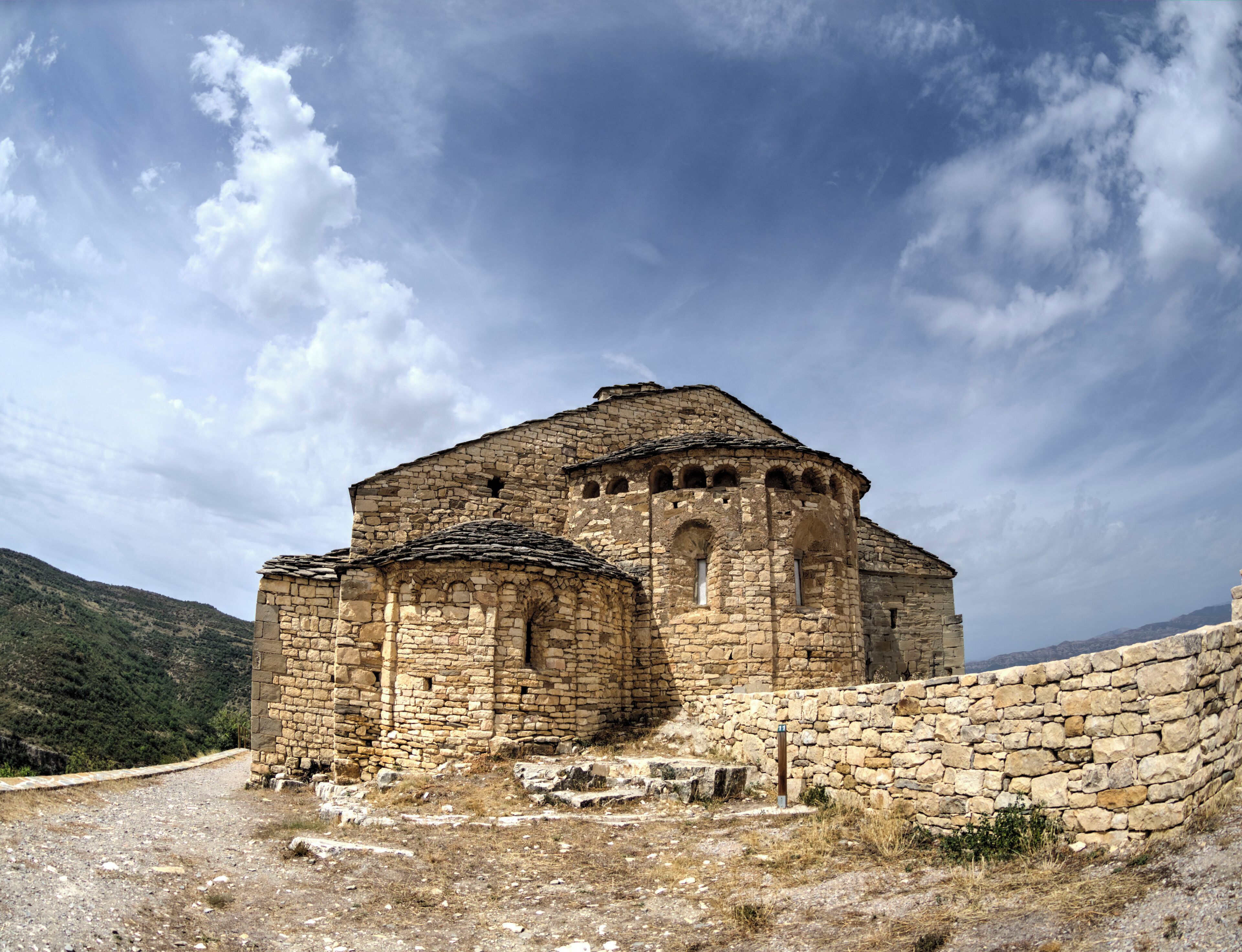 An ancient stone building with a dome and arched windows, surrounded by a stone wall and a mountainous landscape.