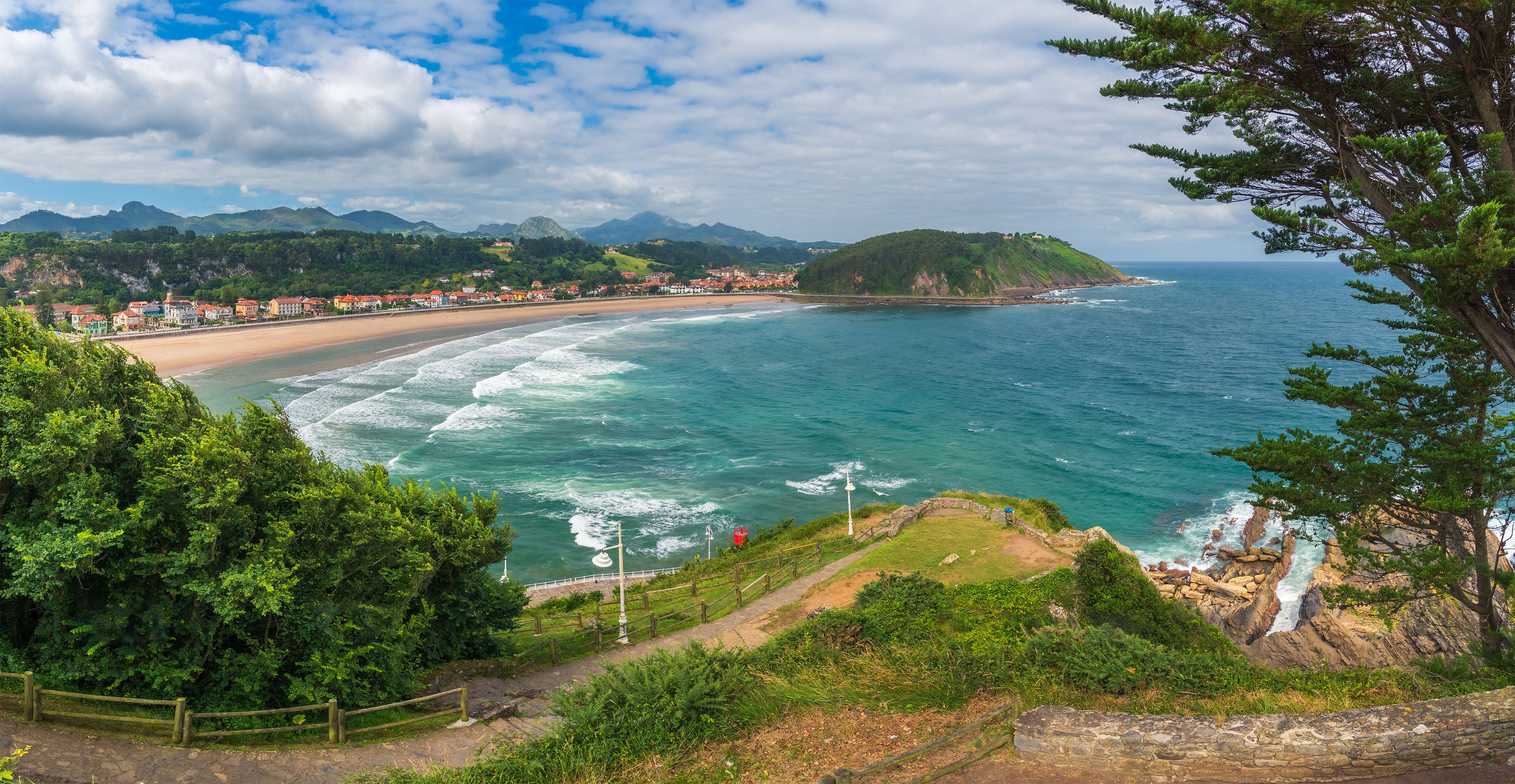 Un paisaje costero con una playa, un pueblo al fondo y montañas a lo lejos.