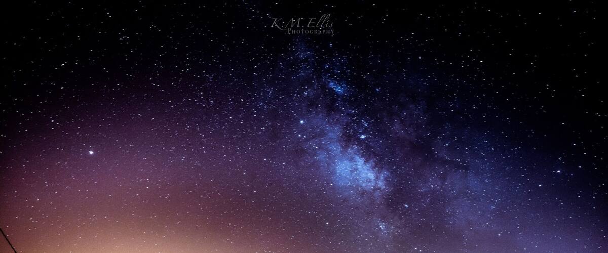 A starry night sky over a landscape with mountains and a tree silhouette.