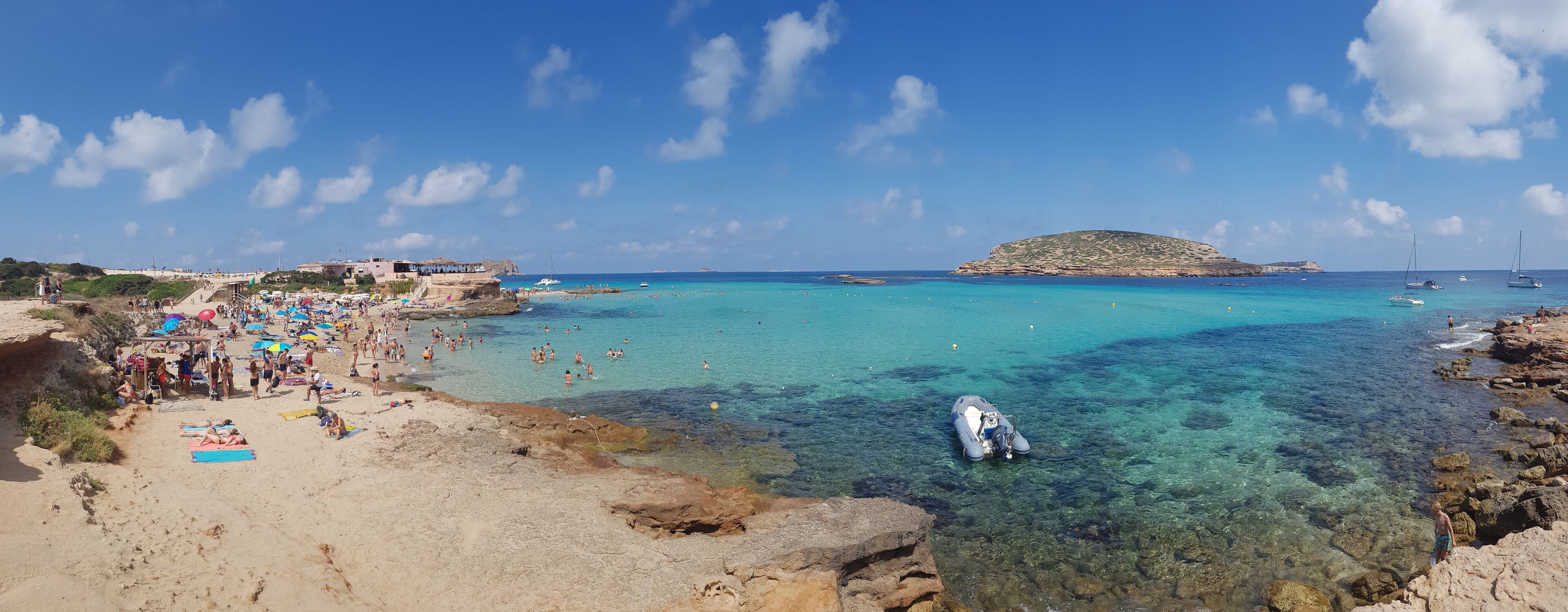 A crowded beach with clear turquoise waters, rocky shorelines, and a small island in the distance.