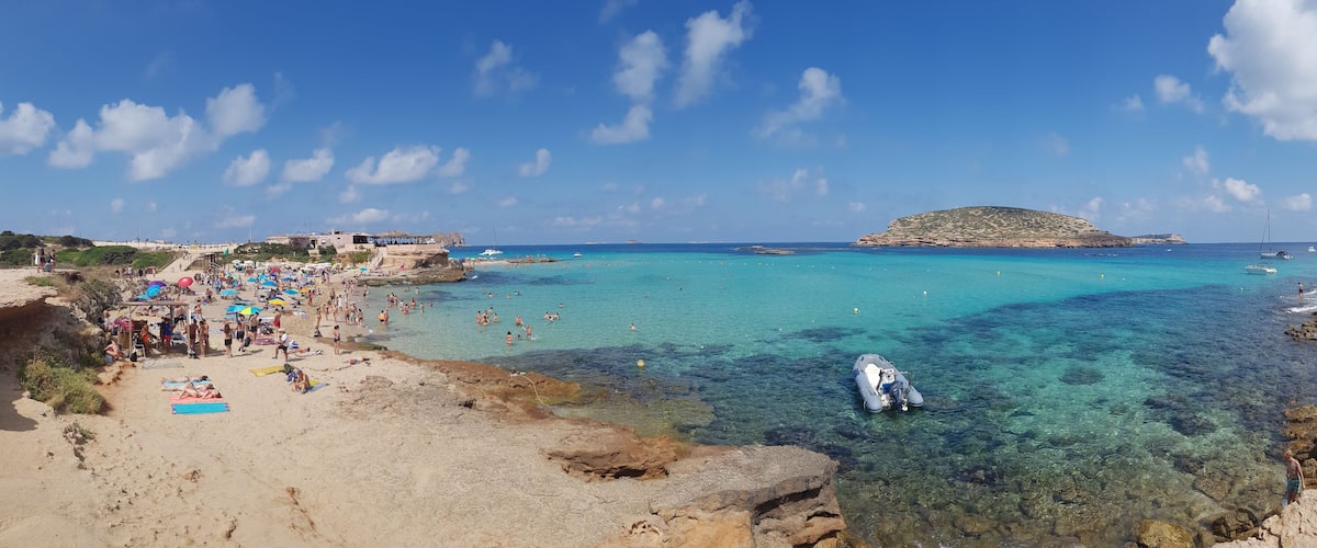 A crowded beach with clear turquoise waters, rocky shorelines, and a small island in the distance.