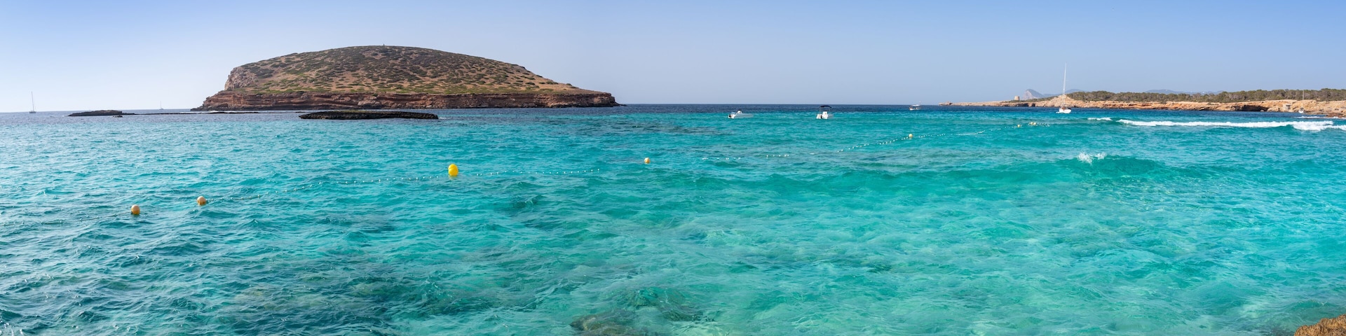 Paisaje costero con aguas turquesas cristalinas, costa rocosa y una pequeña isla con una colina.