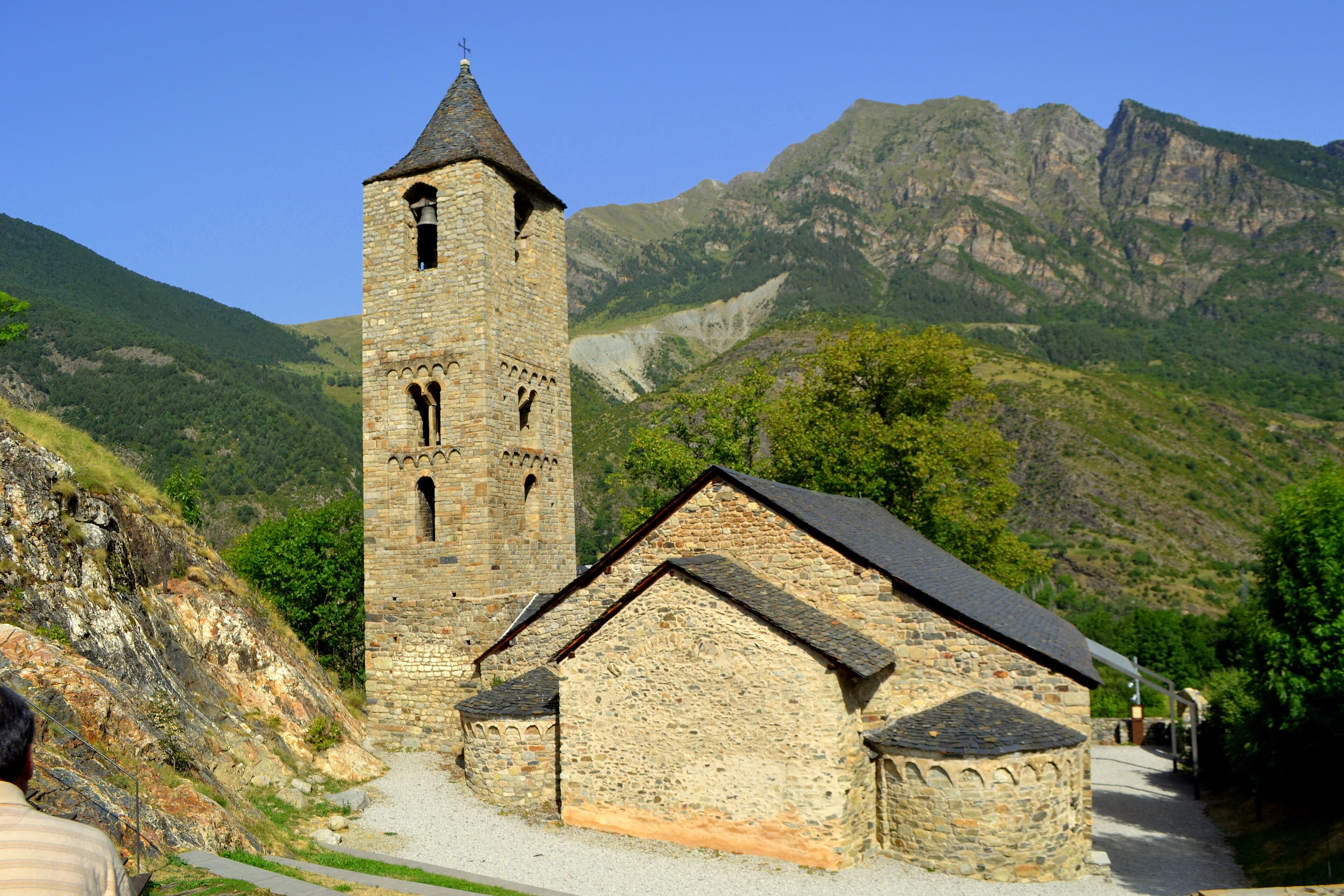 A stone church with a bell tower and a smaller building, surrounded by mountains and greenery.