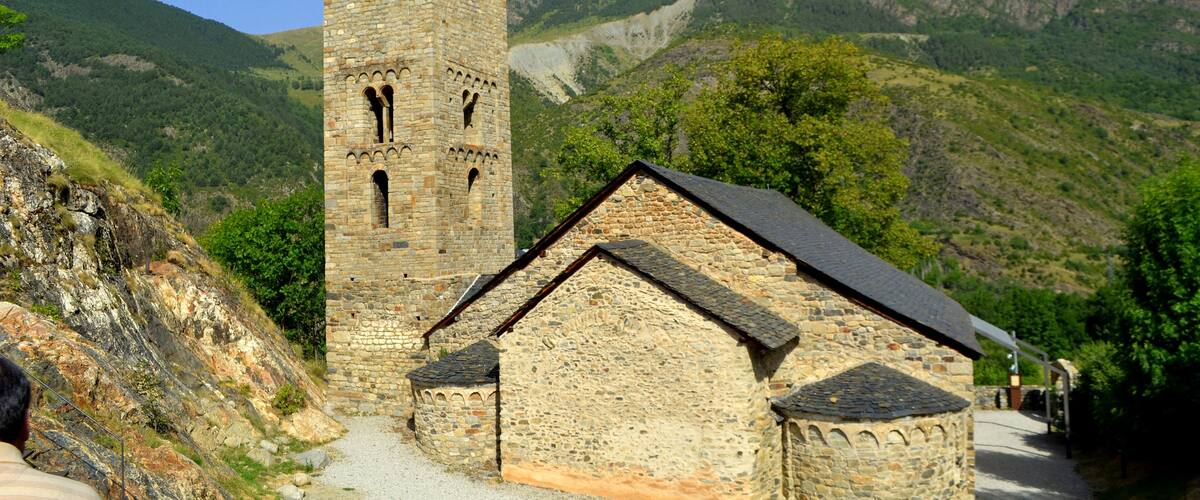 A stone church with a bell tower and a smaller building, surrounded by mountains and greenery.