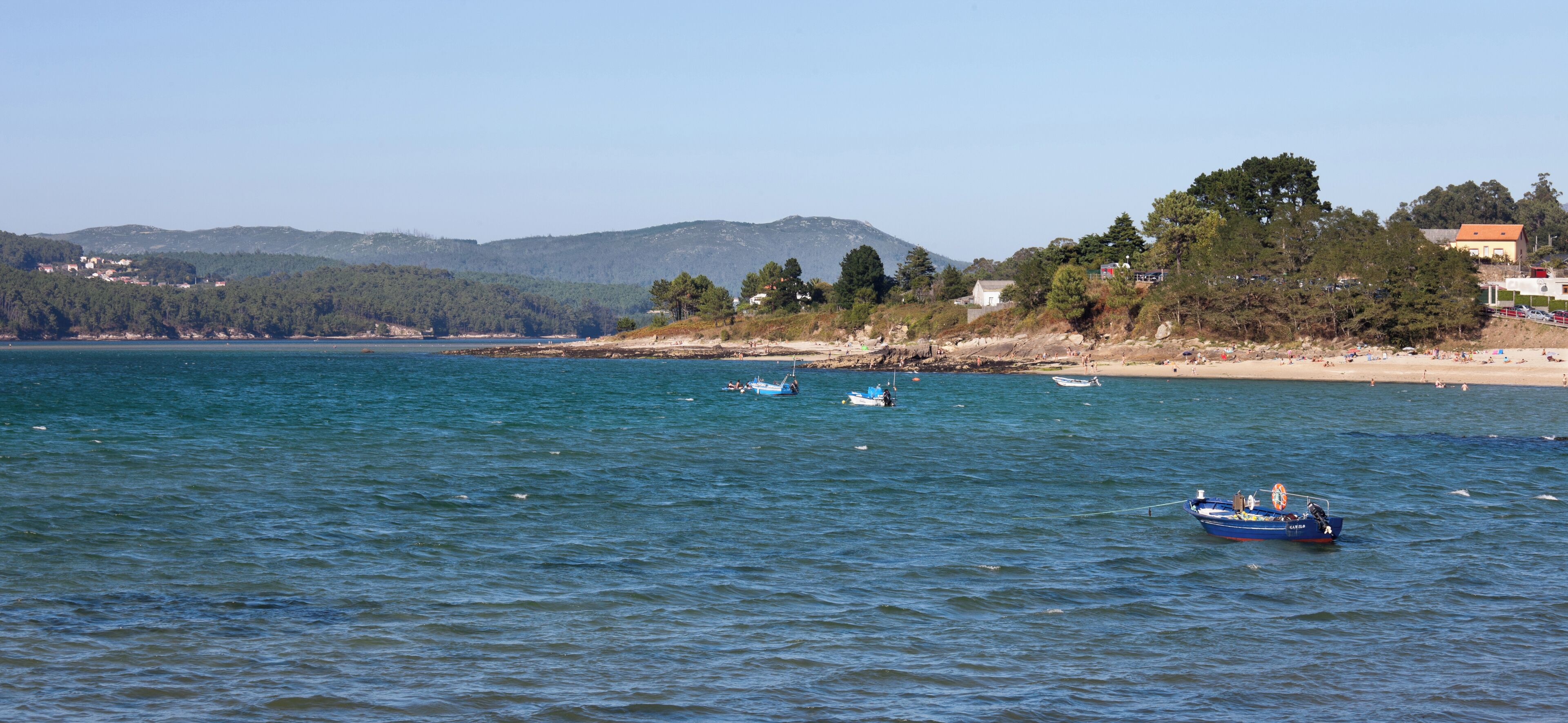 A coastal scene with calm waters, a sandy beach, and a backdrop of hills and trees.