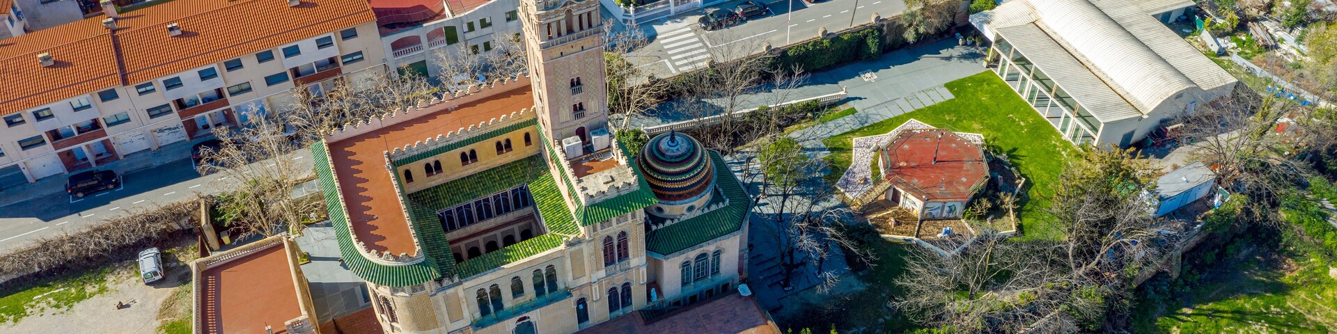 Vista aérea de uma cidade com um edifício histórico imponente, com telhado verde e torre, rodeado por prédios residenciais modernos e uma via principal.