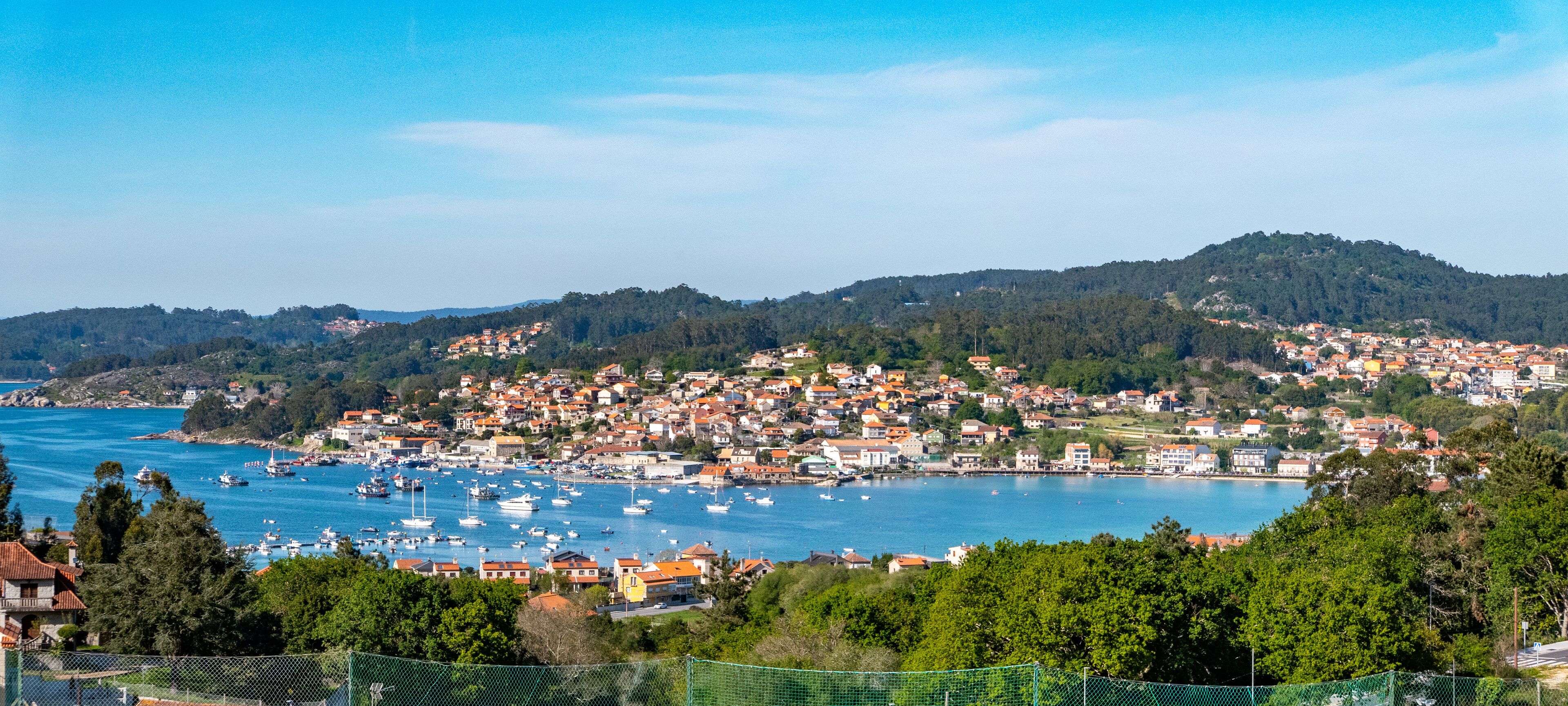 A coastal town with a harbor filled with boats, surrounded by hills and dense greenery.