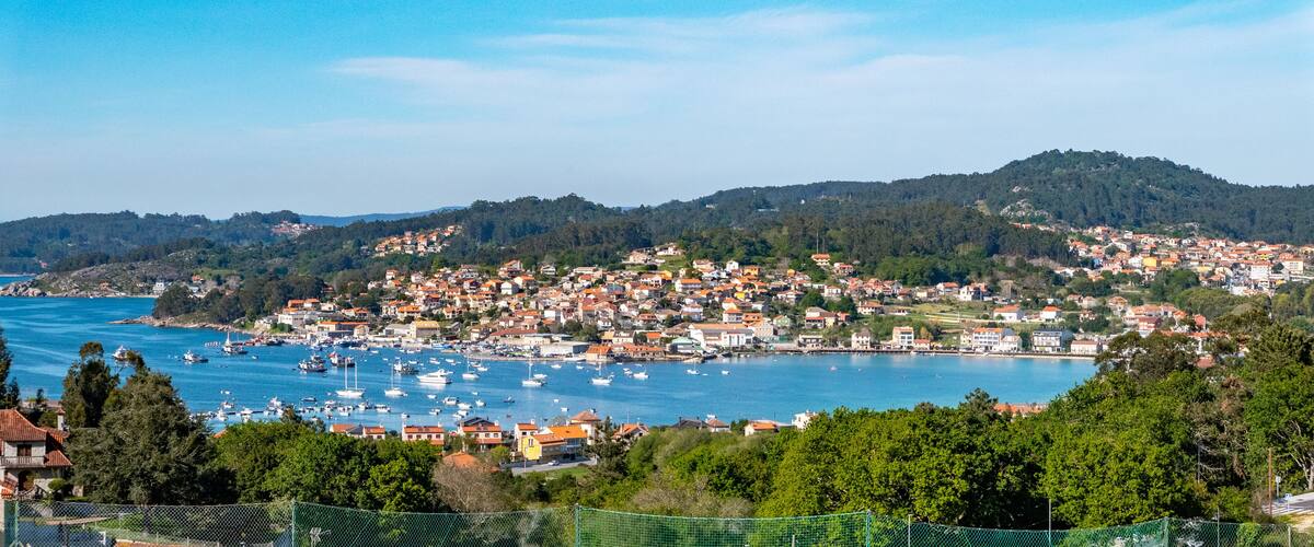 A coastal town with a harbor filled with boats, surrounded by hills and dense greenery.