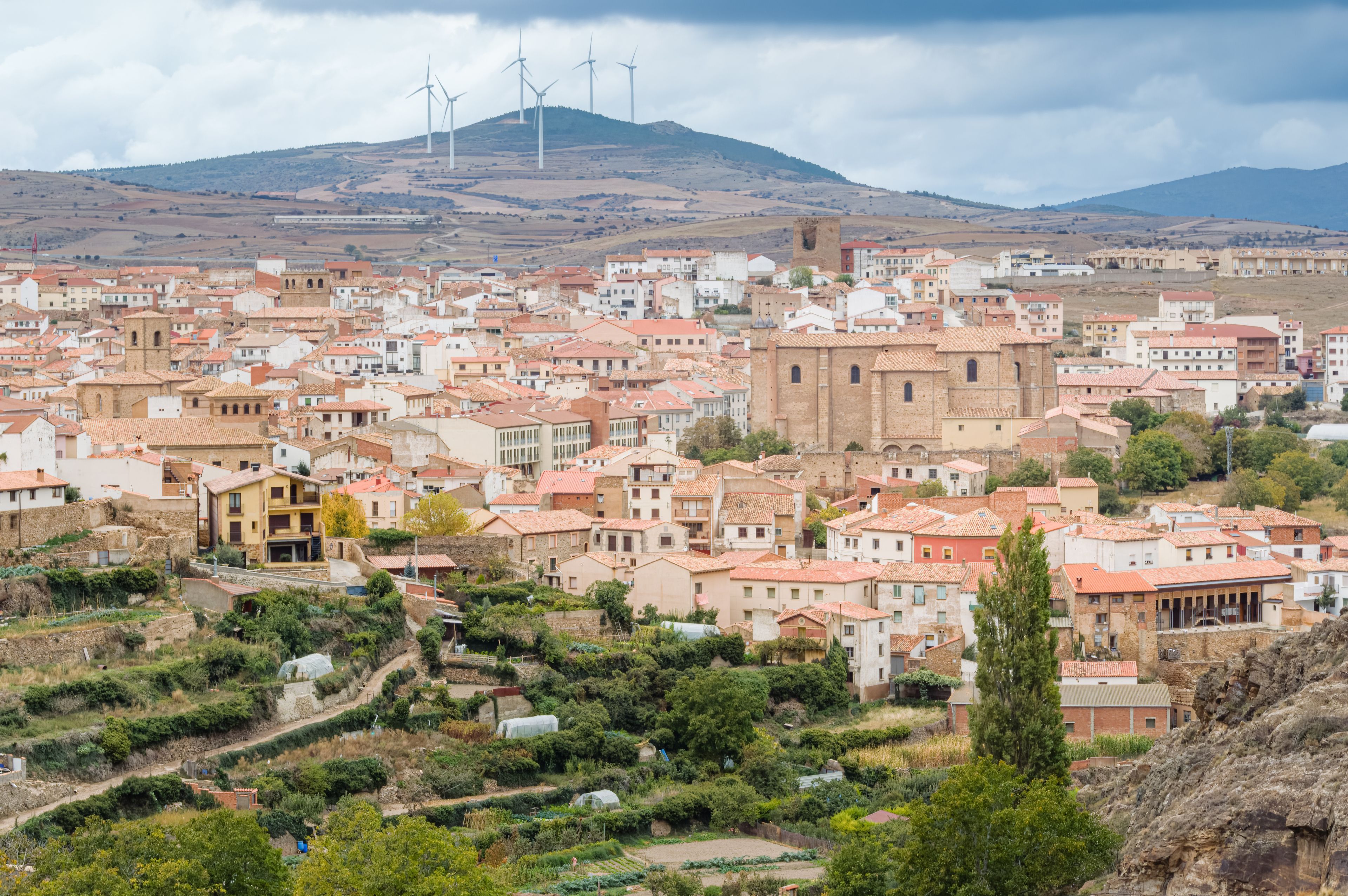 A town with terracotta-roofed buildings, a church, and wind turbines in the background.