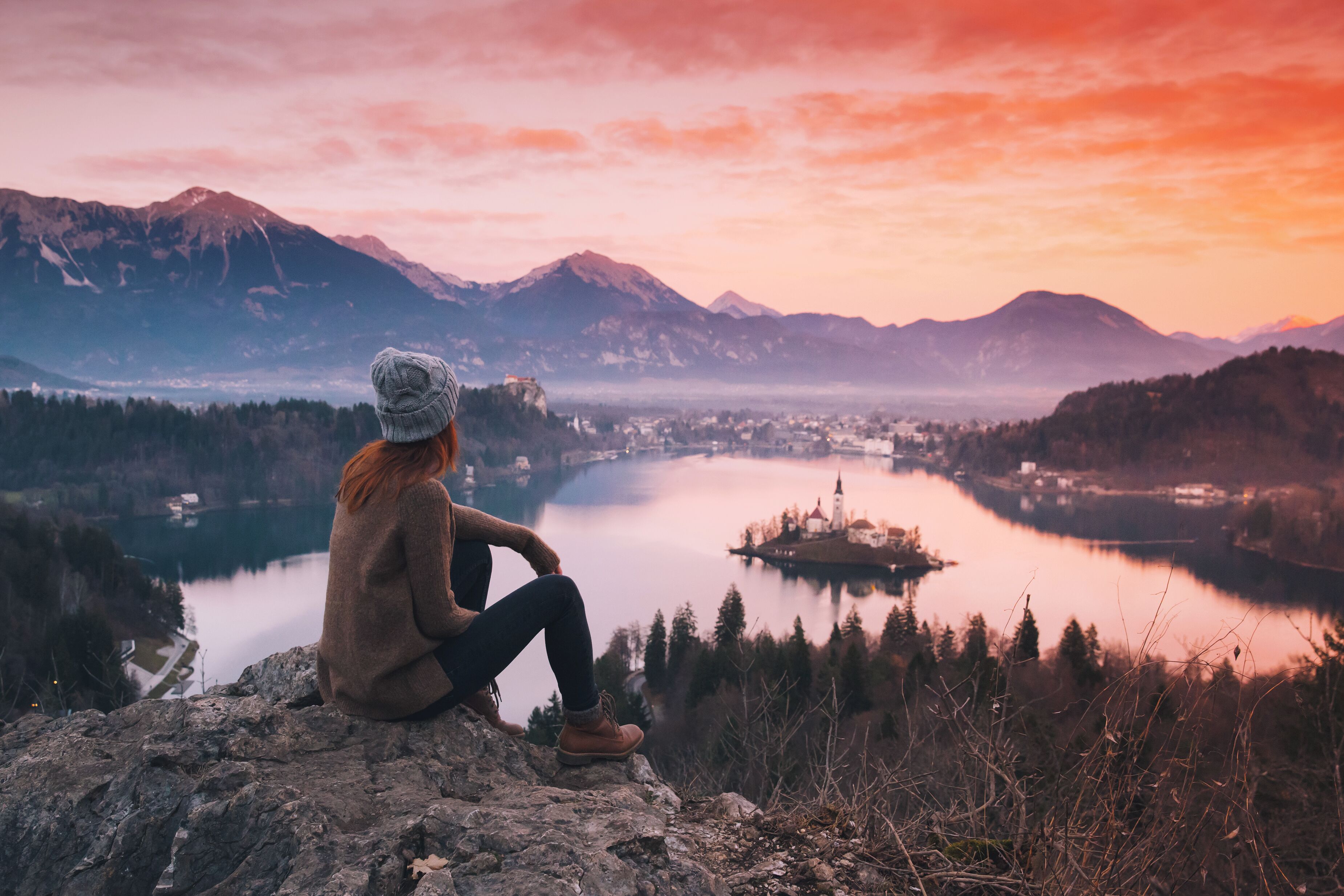 A person sitting on a rocky outcrop overlooking a lake with mountains in the background.