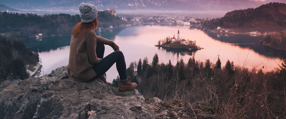 A person sitting on a rocky outcrop overlooking a lake with mountains in the background.