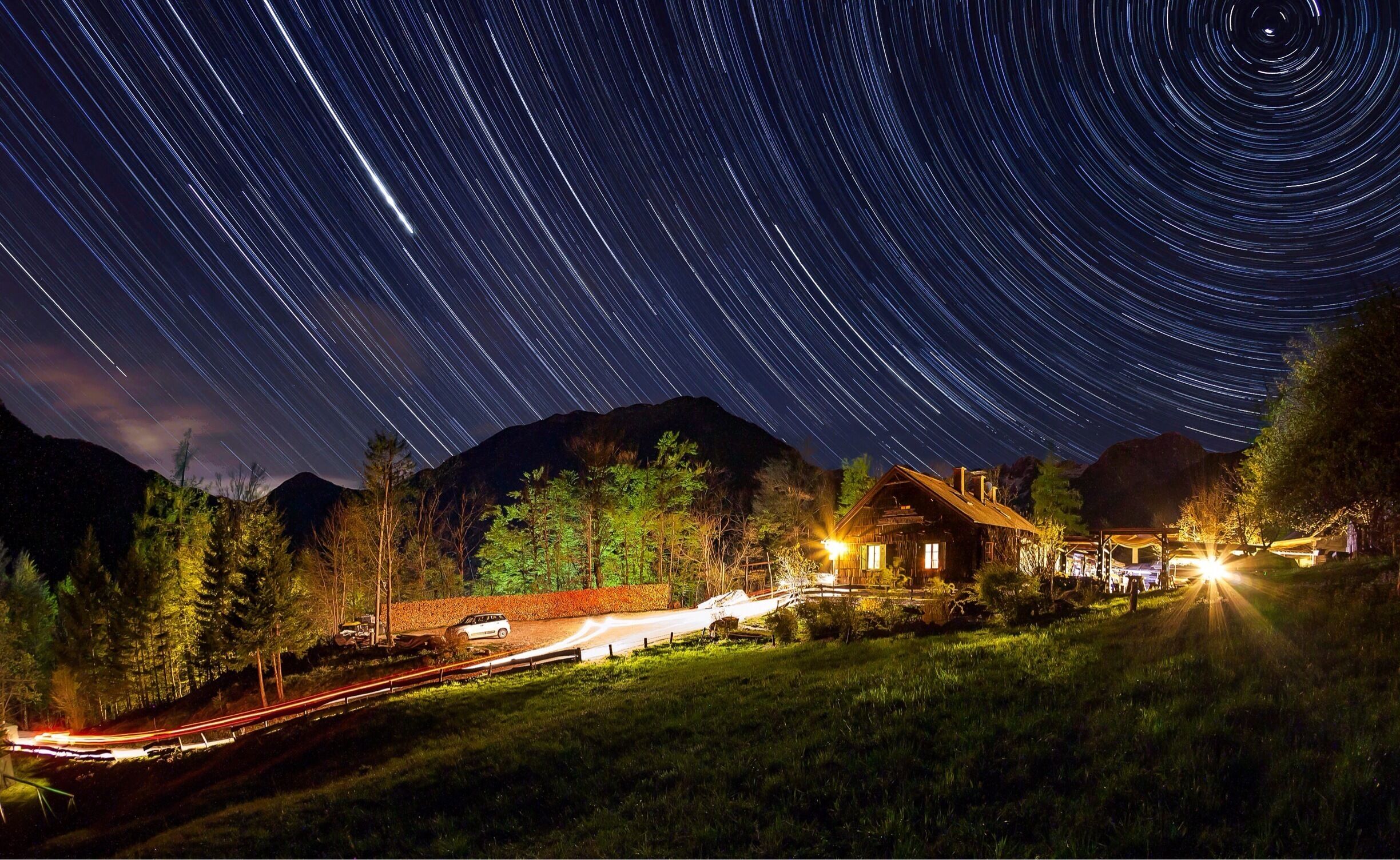 A star trail over a mountain range at night with a lit cabin and a winding road.