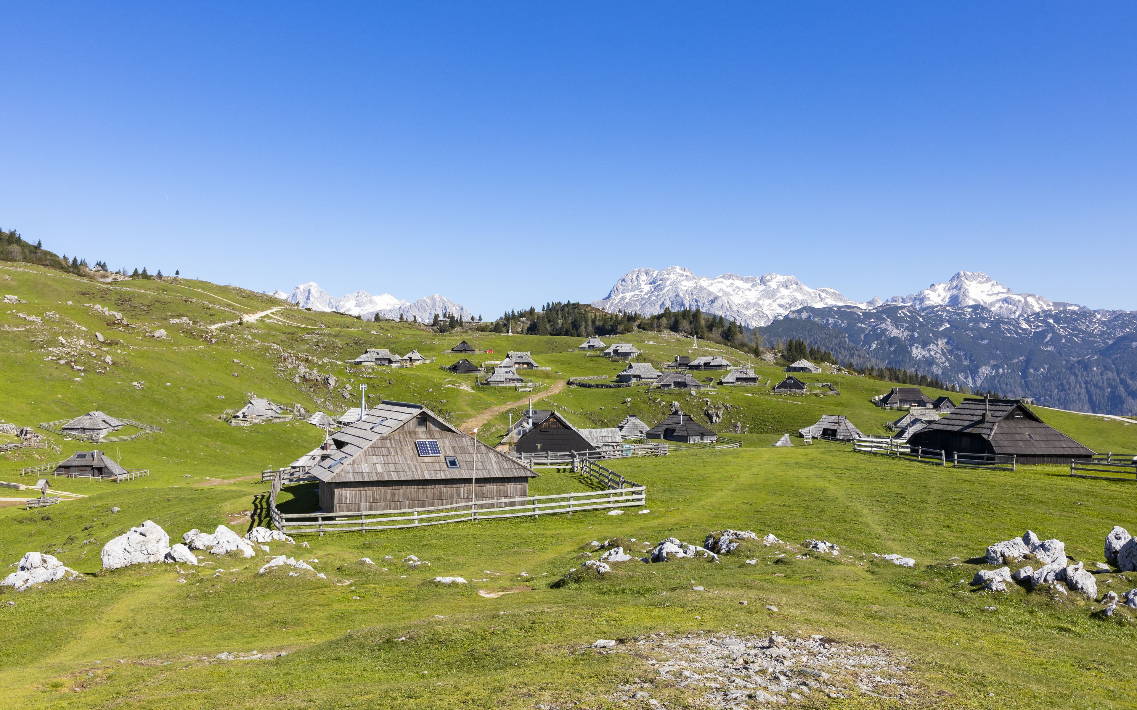 Un village de montagne aux maisons traditionnelles en bois, avec un ciel dégagé et des sommets enneigés en toile de fond.