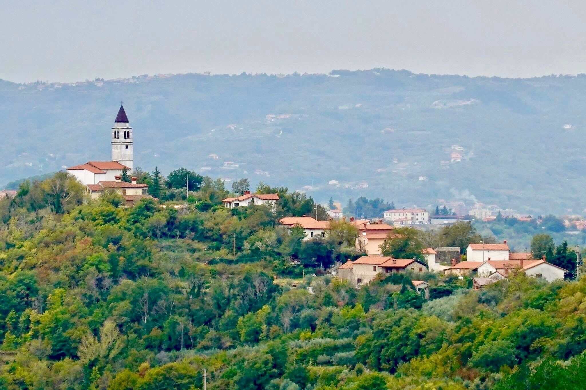 A village with a prominent church tower on a hill, surrounded by lush greenery and scattered houses.