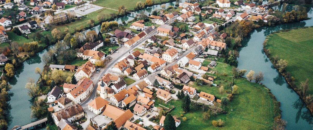 An aerial view of a riverside town with a prominent church, surrounded by fields and a bridge.