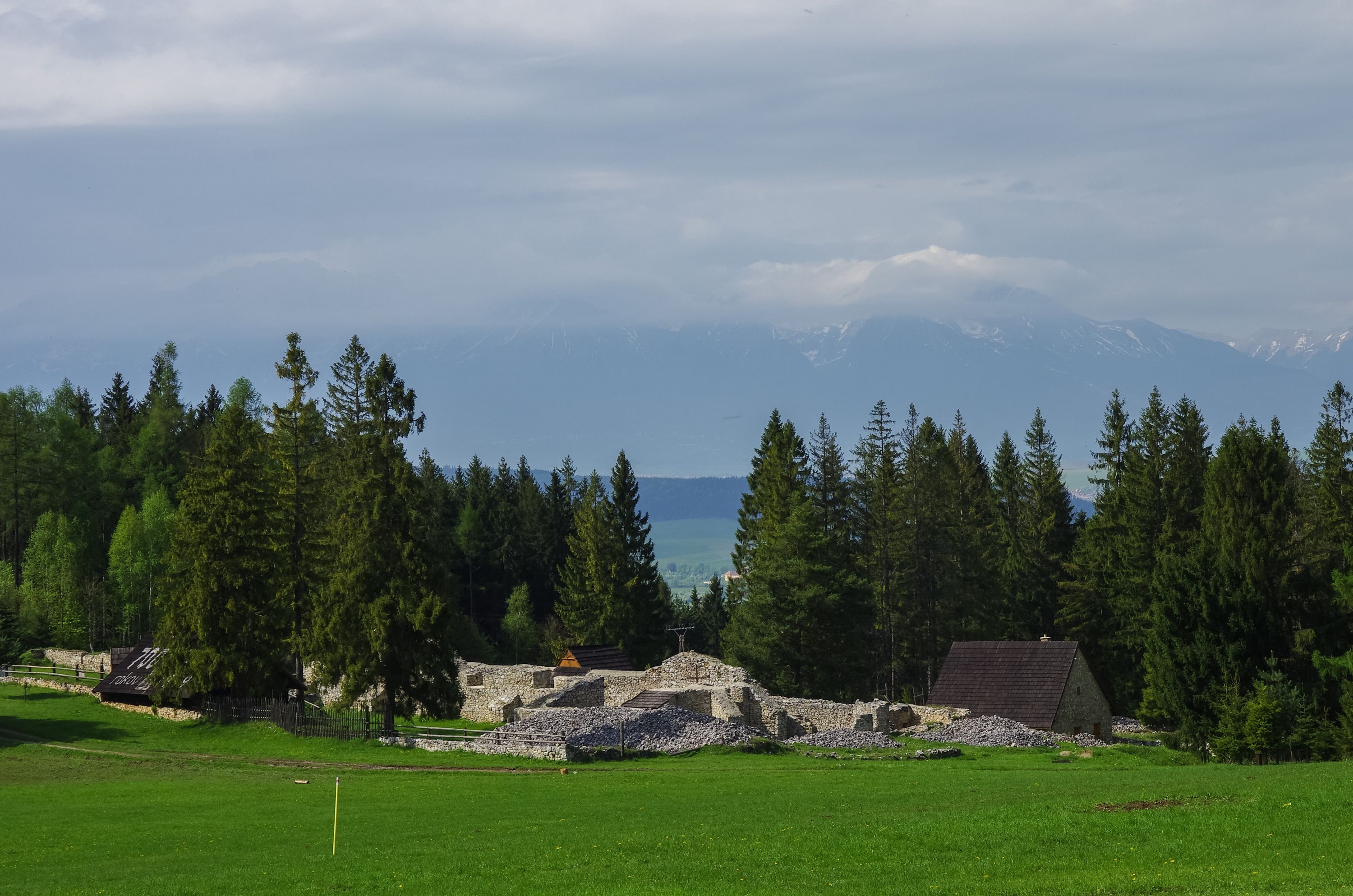 A grassy field with a stone ruin and a wooden structure, surrounded by dense coniferous trees and a mountain range in the background.