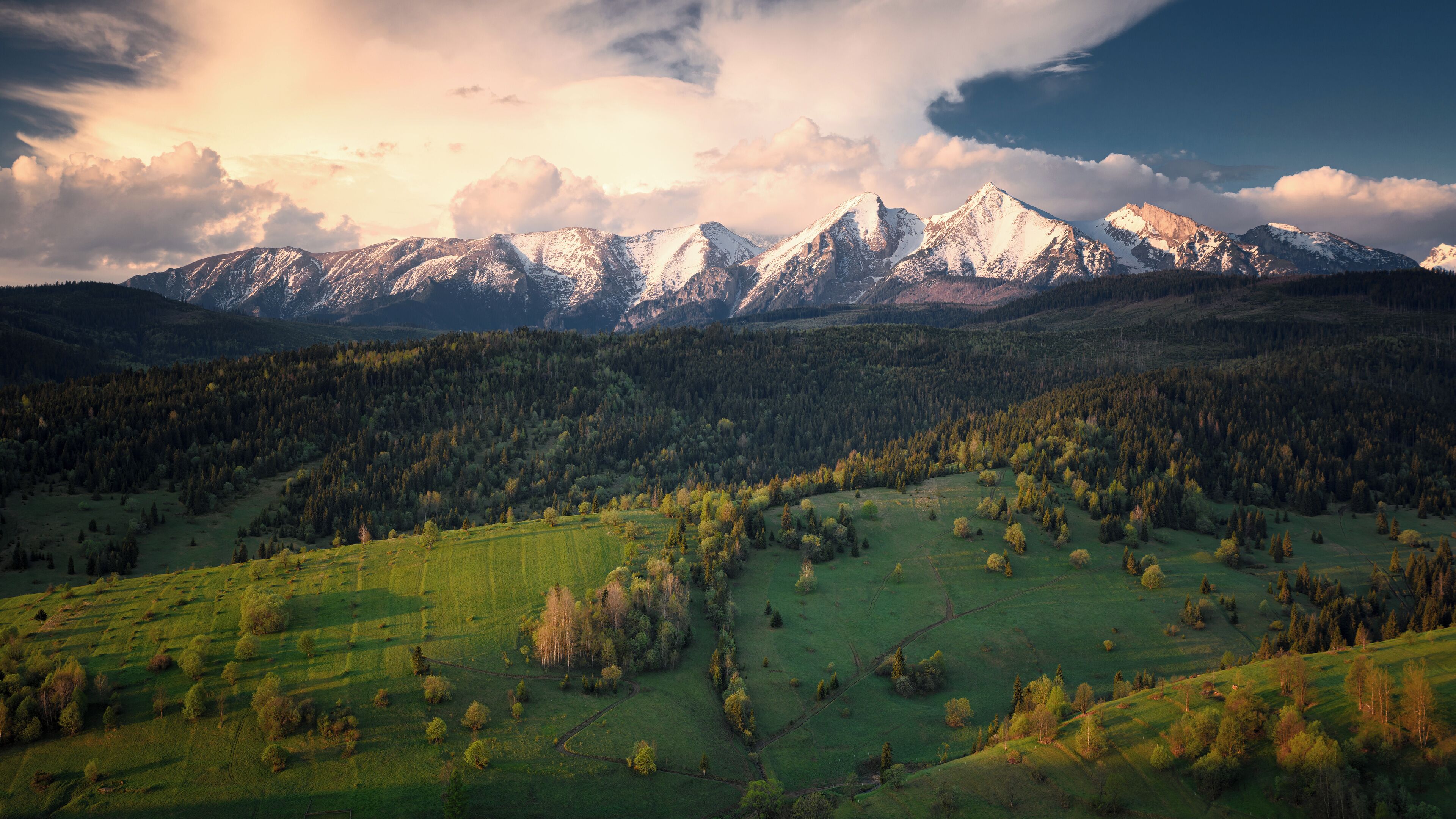 A mountainous landscape with snow-capped peaks, dense forests, and rolling green fields.