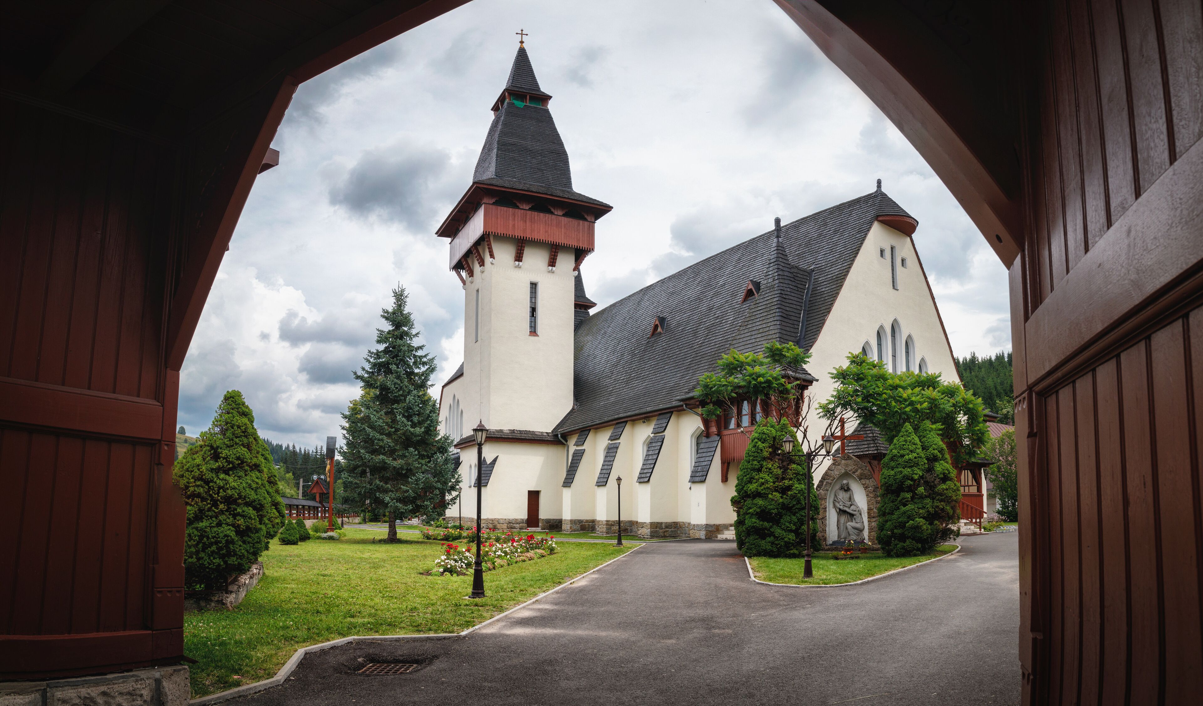 A church with a tall tower and a steep roof, surrounded by greenery and a paved path.
