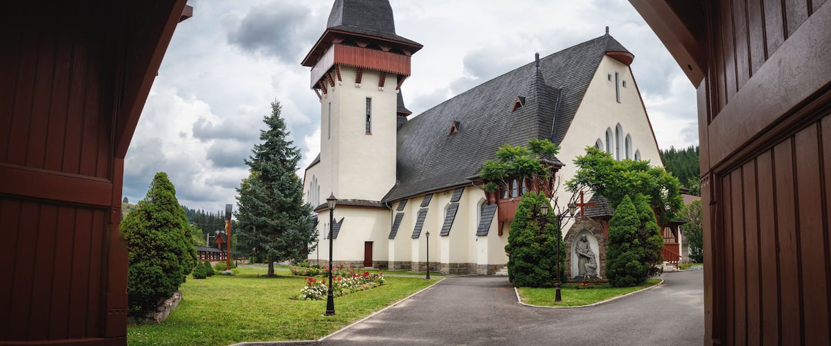 A church with a tall tower and a steep roof, surrounded by greenery and a paved path.