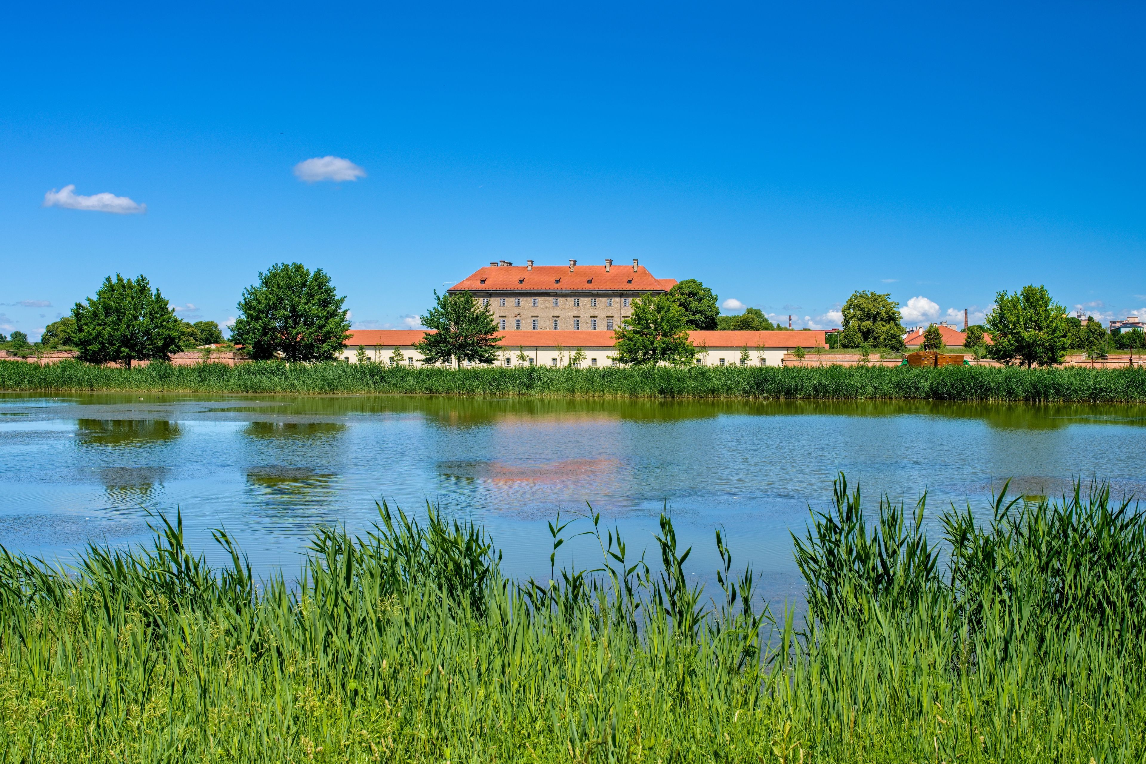 Scenic view of the historical Holic Castle surrounded by green trees and reflected in a peaceful lake under a blue sky in Slovakia.