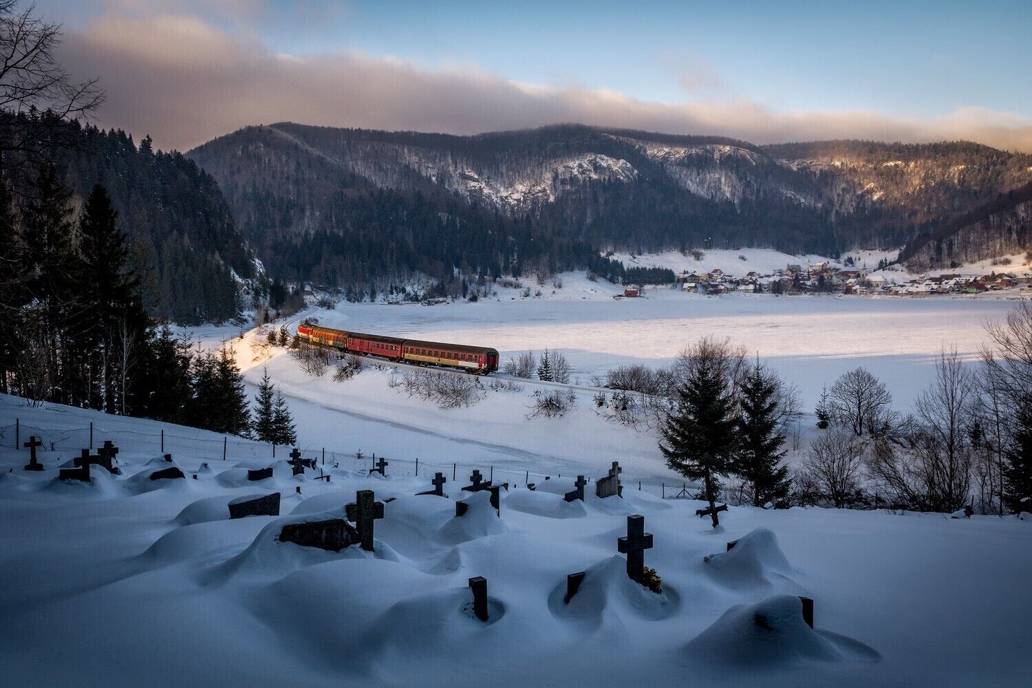 A snow-covered landscape with a train in the distance, a cemetery with crosses, and mountains in the background.