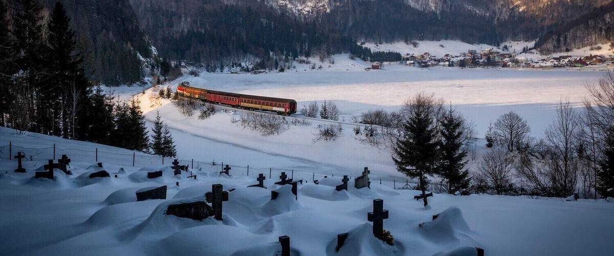 A snow-covered landscape with a train in the distance, a cemetery with crosses, and mountains in the background.