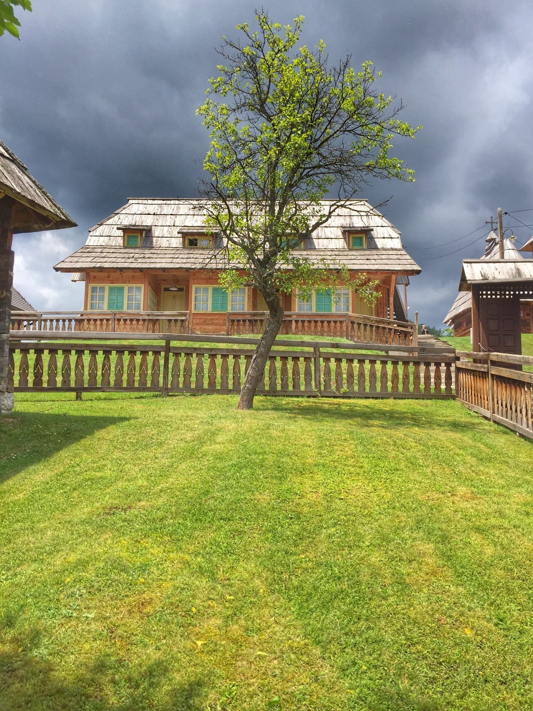 A traditional wooden house with a sloped roof and green shutters, surrounded by a wooden fence and a small tree in the foreground.