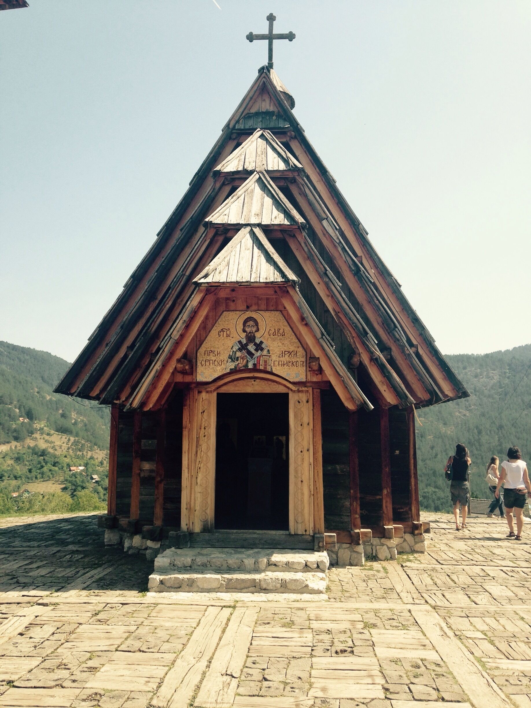 A wooden chapel with a cross on top, set against a mountainous backdrop.