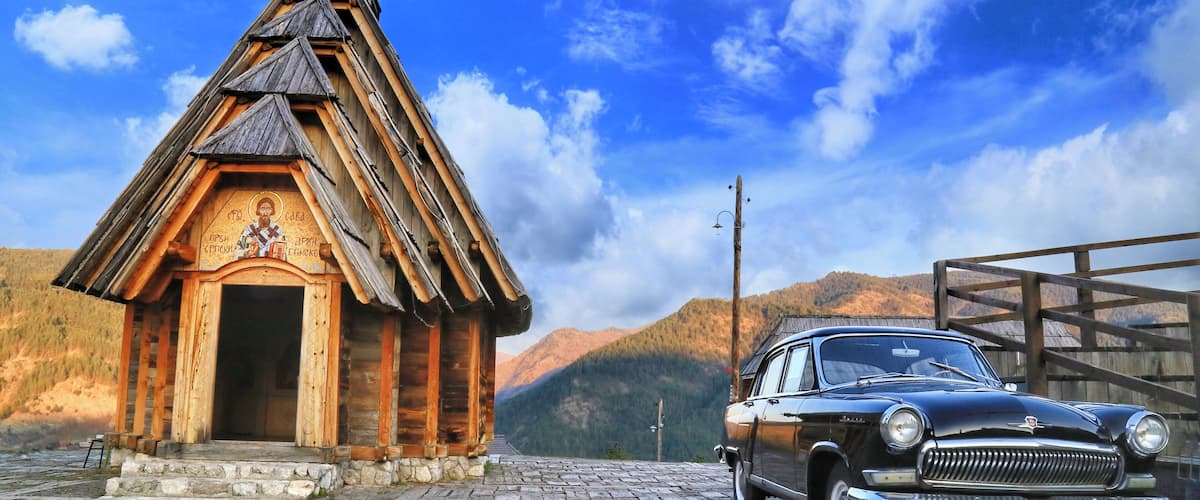 A wooden church with a bell tower, a vintage car parked in front, and mountains in the background.