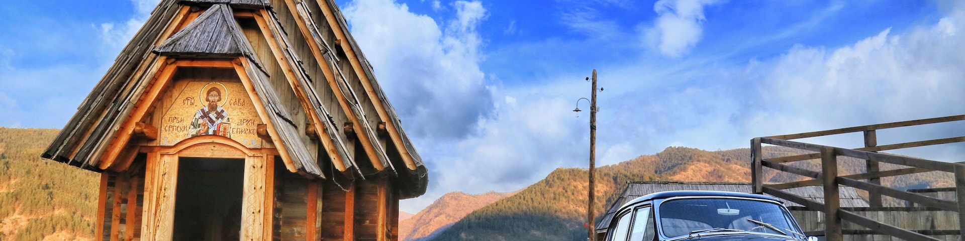 A wooden church with a bell tower, a vintage car parked in front, and mountains in the background.