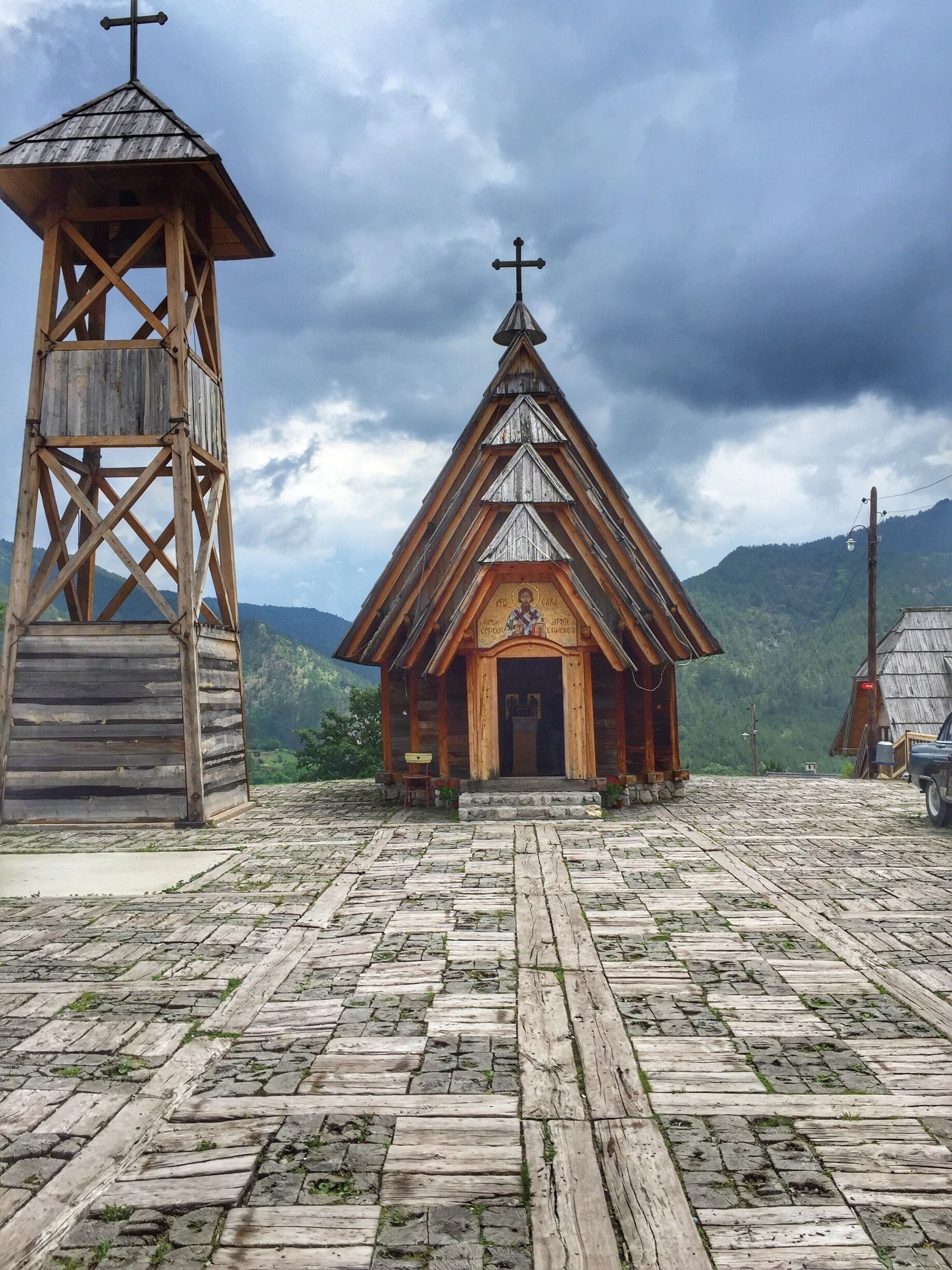A wooden church with a bell tower and a cross on top, surrounded by mountains and a cloudy sky.